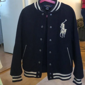 Mörkblå ulljacka från Polo Ralph Lauren - Säljer en mörkblå bomberjacka från Polo Ralph Lauren med vita ränder på muddar och krage. Jackan har knappar framtill, två fickor och en stor broderad logga på bröstet. Klassisk och sportig stil. 