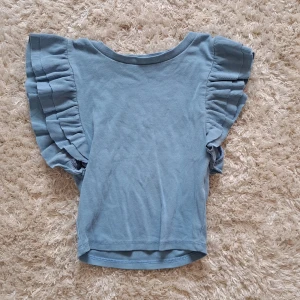 Blå ribbad topp med volangärmar från Zara - Säljer en ljusblå ribbad topp från Zara med korta volangärmar. Toppen har en rund halsringning och är tillverkad i ett mjukt, stretchigt material. Perfekt för en trendig look.