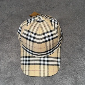 Beige rutig keps från Burberry - Snygg keps från Burberry i klassiskt beige rutigt mönster med svarta, vita och röda detaljer. Kepsen har böjd skärm och justerbar rem baktill. Perfekt accessoar för en trendig look.