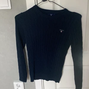 Mörkblå kabelstickad tröja från GANT - Säljer en klassisk mörkblå kabelstickad tröja från GANT i stretchig bomull. Tröjan har rund halsringning, lång ärm och diskret broderad logga på bröstet. Perfekt för en stilren och tidlös look.
