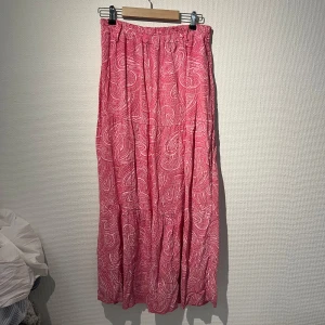 Rosa långkjol med mönster - Säljer en rosa långkjol med vitt paisleymönster. Kjolen har resår i midjan och är luftig med flera volanger. Perfekt för varma dagar och ger en avslappnad men snygg look. Köpt i Spanien💖