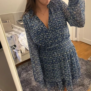 Blå blommig klänning från Zara - Säljer en blå klänning från Zara med ett vackert blommigt mönster. Klänningen har långa ärmar och en smockdetalj i midjan för en fin passform. Perfekt för vår och sommar!