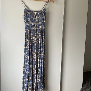 Blommig långklänning från H&M - Säljer en blå och beige långklänning från H&M med smala axelband och knappar framtill. Klänningen har ett blommigt paisleymönster och smockad rygg för extra komfort. Perfekt för varma dagar och somriga tillfällen!