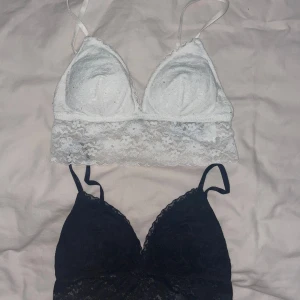 Två spetsiga bralettes i vitt och svart - Säljer två snygga bralettes, en vit och en svart, båda i mjuk spets med smala axelband och lätt vadderade kupor. Perfekta att bära under en skjorta eller som de är till ett par jeans.