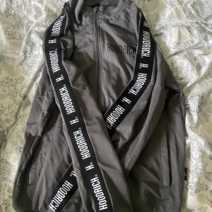 Hoodrich track jacket - Säljer en grå track jacket från Hoodrich med svarta detaljer och tydlig logga längs ärmarna. Jackan har dragkedja framtill och två fickor. Perfekt för en sportig och avslappnad stil. Finns byxor till i en annan annons.