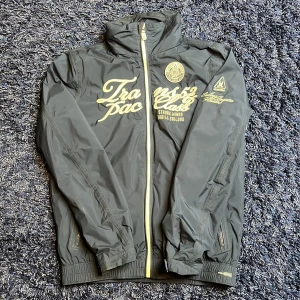 Windbreaker från Gaastra - Säljer en svart vindjacka från Gaastra med vitfärgade broderade loggor och textdetaljer. Jackan har dragkedja framtill, hög krage och ribbade muddar vid ärmslut och nederkant. Perfekt för en sportig och stilren look. Hör av er vid minsta fundering🙌