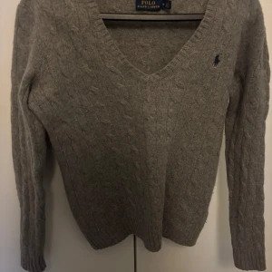 Grå stickad polotröja från Polo Ralph Lauren - Säljer en klassisk grå stickad tröja från Polo Ralph Lauren med v-ringning och kabelstickat mönster. Tröjan har lång ärm och en liten broderad logga på bröstet. Perfekt till jeans eller kjol! Tröjan är tajt i armarna och den kostade ungefär 600kr 