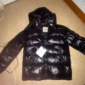 Svart dunjacka från Moncler pris kan diskuteras vid snabb affär😁 - Säljer en svart glansig dunjacka från Moncler med huva och dragkedja. Jackan har en klassisk pufferstil och är perfekt för kyliga dagar. Insidan har ett unikt Moncler-serietryck. Snygg och tidlös design som passar till det mesta.