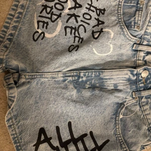 Ljusblå jeansshorts med tryck från FB Sister - Unika ljusblå jeansshorts från FB Sister med svart och vitt tryck framtill och två vita hjärtan på bakfickan. Klassisk femficksmodell med knapp och dragkedja. Perfekta för en cool och personlig stil.