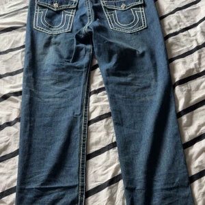Blå jeans från True Religion - Säljer ett par klassiska blå jeans från True Religion med tydliga vita sömmar och snygga bakfickor med lock och knapp. Jeansen har rak passform och normal midja, perfekta för en avslappnad stil. Använda 1 gång