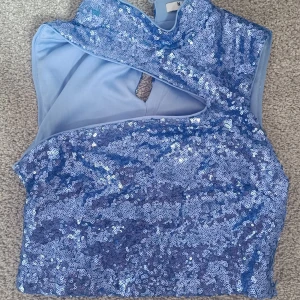 Blå glittrig paljettopp från NA-KD - Säljer en lila glittrig paljettopp från NA-KD i storlek 36. Toppen är  täckt av paljetter för en riktigt festlig look. Stängs med knappar i nacken och har en snygg öppen detalj framtill. Är mer lila i verkligheten. 160 kronor men priset kan diskuteras!! 