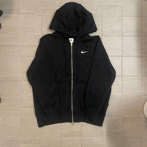 Svart hoodie från Nike - Selling a black hoodie from Nike with a zipper and hood. It has a white Nike logo on the chest and practical pockets at the front. Perfect for a relaxed style. Has only been used twice. Too big in size, original price 90$. It doesn't say that it's large in size, but it is.