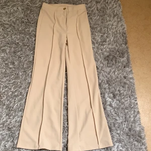 Beiga kostymbyxor med pressveck - Snygga beiga kostymbyxor med hög midja och vida ben. Byxorna har pressveck framtill och stängs med knapp och dragkedja. Perfekta för en stilren look.