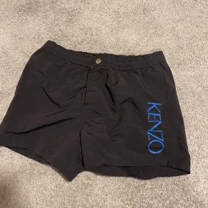 Svarta bad shorts från Kenzo med blå logga - Svarta shorts från Kenzo med blå logotyp längs ena benet. Shortsen har elastisk midja med snörning, knapp och innerfoder i mesh. Perfekta för sommaren och har en sportig look.
