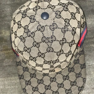 Gucci keps - Säljer en beige keps från Gucci med klassiskt GG-monogram i svart över hela kepsen. Kepsen har justerbar rem bak i svart läder och röda/blå detaljer på sidorna. Perfekt accessoar för en trendig look. Använd få gånger, har bytt stil därför säljer jag den. Det är en blue canavas men den ser beige ut med blixt