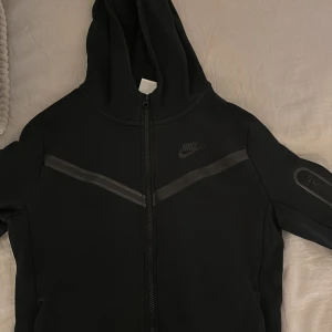 Svart hoodiejacka från Nike (Nike tech) - Svart hoodiejacka från Nike med dragkedja framtill och huva. Jackan har diskreta reflexdetaljer på bröstet och ärmen samt Nike-logga på bröstet. Perfekt för en sportig och avslappnad stil. Detta är en junior storlek så den passar både S/XS