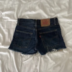 Levi's jeansshorts  - Mörkblå jeansshorts från Levi's med fransig, rå kant nertill. Klassisk femficksmodell med knapp- och dragkedjegylf. Levi's läderpatch bak i midjan och synliga sömmar. Perfekta för en avslappnad stil.