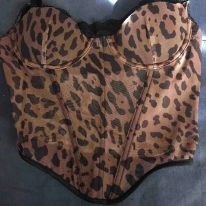 Leopardmönstrad korsett med spetsdetaljer - Säljer en snygg leopardmönstrad korsett med svarta spetsdetaljer längs kanten. Modellen har bygel och ger bra stöd, perfekt för att lyfta din outfit med lite extra edge.