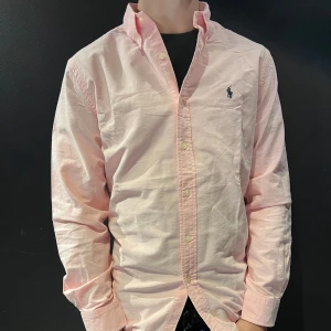 Rosa skjorta från Ralph Lauren - Säljer en klassisk rosa skjorta från Ralph Lauren med broderad logga på bröstet. Skjortan har lång ärm, knappar framtill och en normal passform. Perfekt till jeans eller chinos för en clean look.