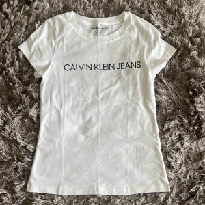 Vit t-shirt från Calvin Klein Jeans - Vit t-shirt från Calvin Klein Jeans med svart logotyptryck över bröstet. Modellen är slim fit med rund halsringning och korta ärmar. Perfekt basplagg med stilren design. Aldrig använd, ny med prislapp