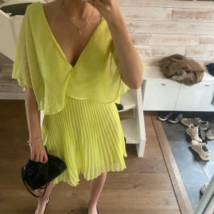 Ljusgul plisserad klänning med volang - Superfin ljusgul klänning med plisserad kjol och volangig överdel. Klänningen är kort och har öppen rygg med bred axelband. Perfekt för sommarens tillfällen när du vill sticka ut lite extra.