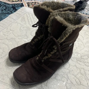 Bruna fodrade boots med snörning - Säljer ett par bruna boots med snörning och mjukt, lurvigt foder på insidan. Skorna har en rund tå och är perfekta för kyligare dagar. Yttermaterialet är mocka och sulan är svart och mönstrad för bra grepp.