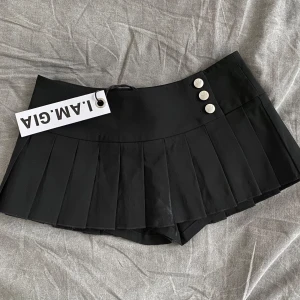 Svart plisserad kjol från I.AM.GIA - Supersnygg svart plisserad kortkjol från I.AM.GIA med tre silvriga knappar framtill. Kjolen har en klassisk tennisstil och är perfekt för att skapa en trendig look. Råkade bränna dem me stryckgärnet framtil, därav priset (går att se i sista bilden)