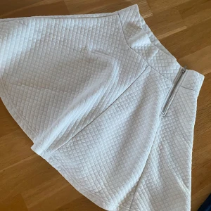 Vit quiltad kjol med dragkedja  - Hej! Min kompis säljer en vit quiltad kjol med markerad dragkedja framtill. Kjolen har en A-linjeformad siluett och är tillverkad i ett mjukt, vadderat material med rutmönster. Perfekt för dig som vill ha en stilren och trendig look.