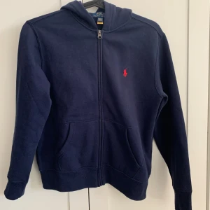 Mörkblå hoodie från Polo Ralph Lauren Barn - Barnstorlek! Mörkblå hoodie med dragkedja från Polo Ralph Lauren. Klassisk design med huva, två fickor framtill och röd broderad logga på bröstet. Perfekt för en avslappnad stil.