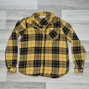 Gul och svart rutig skjorta från H&M - Snygg gul och svart rutig skjorta från H&M i storlek 158. Skjortan har långa ärmar, klassisk krage och knäppning framtill. Bröstficka med rutigt mönster på snedden för extra detalj. Perfekt för en cool och avslappnad stil.
