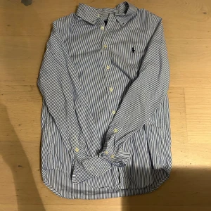 Randig skjorta från Ralph Lauren - Klassisk blå- och vitrandig skjorta från Ralph Lauren med lång ärm och knappar framtill. Skjortan har en broderad logga på bröstet och är tillverkad i mjuk bomull. Perfekt för en clean och stilren look.