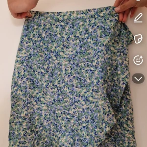 Blommig omlottkjol med volang - Söt omlottkjol med blått, grönt och vitt blommönster. Kjolen har volangkant nertill och knyts i midjan för en snygg passform. Har inga skador och har använts ett fåtal gånger. 