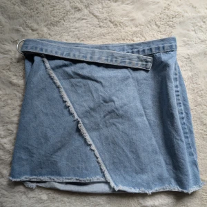 Ljusblå omlottkjol i jeans - Trendig ljusblå jeanskjol med omlottmodell och råa kanter. Kjolen har ett bälte i midjan med metallring och cool fransig detalj framtill. Perfekt för dig som gillar en avslappnad och edgy stil.