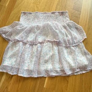 Söt blommig volangkjol i pastell - Pastellfärgad kortkjol med småblommigt mönster i ljusblått och rosa. Kjolen har bred smock i midjan och dubbla volanger för extra flowy känsla. Perfekt för varma dagar och ger en romantisk vibe.