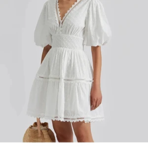 Vit kortklänning med puffärm By Malina - Superfin vit kortklänning från By Malina med puffiga korta ärmar, markerad smock i midjan och v-ringning med spetsdetaljer. Klänningen har diskret prickigt mönster och volangkant nertill. Perfekt för sommarens alla tillfällen! Endast använd en gång! Nypris: 2400