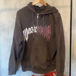 Brun hoodie från Wasted Paris - Säljer en brun hoodie från Wasted Paris med vit och rosa tryckt text på bröstet. Tröjan har dragkedja, huva med snörning och stora fickor framtill. Perfekt för dig som gillar streetwear och vill ha en chill look. Pris kan diskuteras 