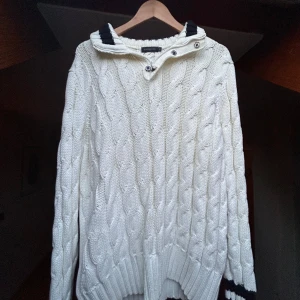 Vit kabelstickad tröja Henri Lloyd - Vit kabelstickad tröja från Henri Lloyd med hög krage och knappar. Tröjan har marinblå detaljer vid kragen och ärmslut samt ett broderat märke på ena ärmen. Perfekt för dig som gillar klassisk och nautisk stil. Lite oversized i storleken. Perfekt nu i varma vädret.    Nyskick. Inte använd alls. Ny pris 2 600, pris kan diskuteras
