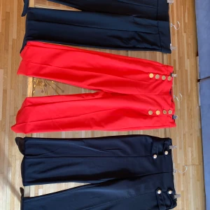 Färgglada kostymbyxor med knappar - Säljer flera par kostymbyxor i olika färger: svart, röd, vinröd, blå och beige. Byxorna har raka ben, hög midja och dekorativa guldknappar längs sidorna. Snygg pressveck framtill och tillverkade i ett slätt, vävt material som ger en dressad look. 1 för 125 och alla tillsammans för 600. Storlek Xs men passar S också