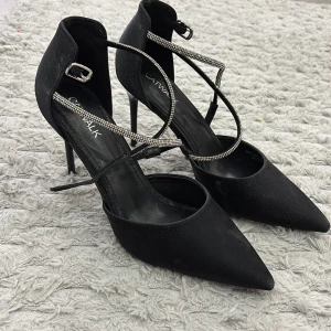 Svarta pumps med glittriga remmar Catwalk - Snygga svarta pumps från Catwalk med spetsig tå och höga klackar. Skorna har glittriga remmar som korsar över foten och ger en extra touch. Materialet är matt och mockaliknande, vilket gör dem stilrena och trendiga.
