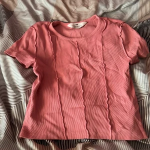 Rosa ribbad topp från H&M - Säljer en rosa ribbad topp från H&M med korta ärmar och rund halsringning. Toppen har dekorativa vågiga sömmar framtill som ger en cool detalj. Perfekt för dig som gillar en enkel men trendig stil.