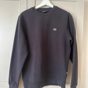 Svart sweatshirt från Dickies - En stilren svart sweatshirt från Dickies med rund halsringning och liten logotyp på bröstet. Tröjan har långa ärmar och är tillverkad i mjuk bomullsmix som är perfekt för lager-på-lager. Enkel och clean look som funkar till det mesta.