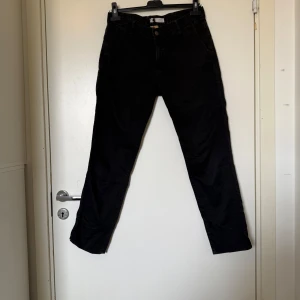 Svarta chinos med tapered fit - Svarta chinos med tapered passform och klassiska fickor fram och bak. Byxorna har bälteshällor och stängs med knapp och dragkedja. Materialet är mjukt och känns som bomull, perfekt för en clean och stilren look. Från märket Odd Molly i storlek 1 som ungefär motsvarar en storlek S. Fint skick, men det är blekare än nya. Och de kan vara rymliga i storlek. 