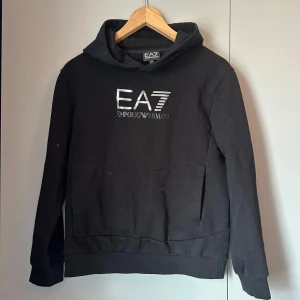 Svart EA7 hoodie med logga - Svart hoodie från EA7 Emporio Armani med stor silvrig logga framtill. Klassisk huva, känguruficka och ribbade muddar. Perfekt för dig som gillar stilrena och exklusiva streetwear-plagg.