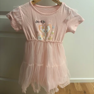 Söt rosa tyllklänning med kattmotiv - Supersöt ljusrosa kortklänning med volangärmar och tyllkjol. Framsidan har ett gulligt kattmotiv och små tryckta detaljer. Överdelen är i mjuk bomull och kjolen i flera lager tyll för extra fluff. Perfekt för dig som älskar söta och lekfulla plagg!