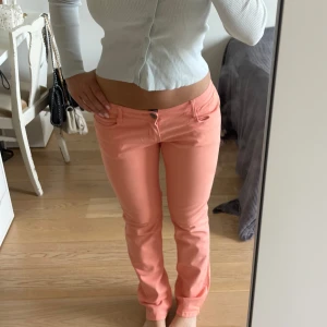 Lågmidjade korallrosa jeans - Snygga lågmidjade jeans i korallrosa från MoD med unika, detaljerade bakfickor. Byxorna är vintage så de finns inte att köpa längre. Färgen är helt perfekt och materialet är super skönt så de sitter jättebra. Skulle säga att modellen är mer bootcut än straight och de är i storlek 30/32, passar mig som vanligtvis har M/38. Fråga gärna om mått!