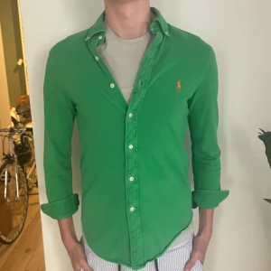 Grön skjorta från Ralph Lauren  - Snygg grön skjorta från Ralph Lauren i storlek S. Klassisk button-down med orange broderad logga på bröstet. Tillverkad i lätt bomullsmaterial, perfekt för dig som gillar stilrena och färgstarka plagg. Långärmad och med knappar hela vägen.