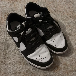 Nike Dunk Low svart/vit sneakers - Nike Dunk Low sneakers i klassisk svart och vit färg. Skorna har svart Swoosh, svarta snören och paneler i svart skinn, medan resten är vitt skinn. Perforerad tåbox och låg profil ger en clean och tidlös look. Perfekta för dig som gillar streetwear och sneakers.