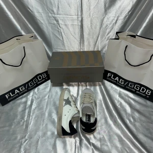Golden Goose vita sneakers med glitterstjärna - Golden Goose sneakers i vitt skinn med silverglittrig stjärna på sidan och svart glitterdetalj på hälen. Grå snörning och beige sula ger en cool kontrast. Märket syns tydligt på häl och plös. Perfekta för dig som gillar statement-detaljer.