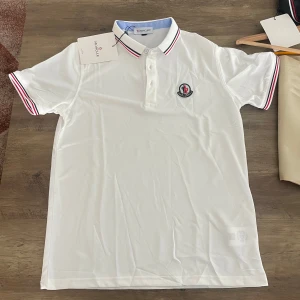 Vit Moncler pikétröja  - Snygg vit pikétröja från Moncler med klassisk krage och knäppning framtill. Tröjan har röda och marinblå ränder på krage och ärmslut samt Moncler-logga broderad på bröstet. Tillverkad i bomull för en fräsch och bekväm känsla.