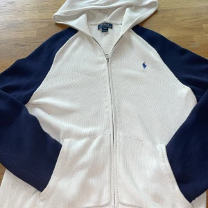 Vit och marinblå hoodie från Polo Ralph Lauren - En vit och marinblå zip hoodie ifrån polo Ralph Lauren tyvärr saknas qr koden men är äkta skick skulle jag säga är 8/10 använd 3 gånger så letar ny ägare priset inte hugget i stenen bara skriva vid funderingar